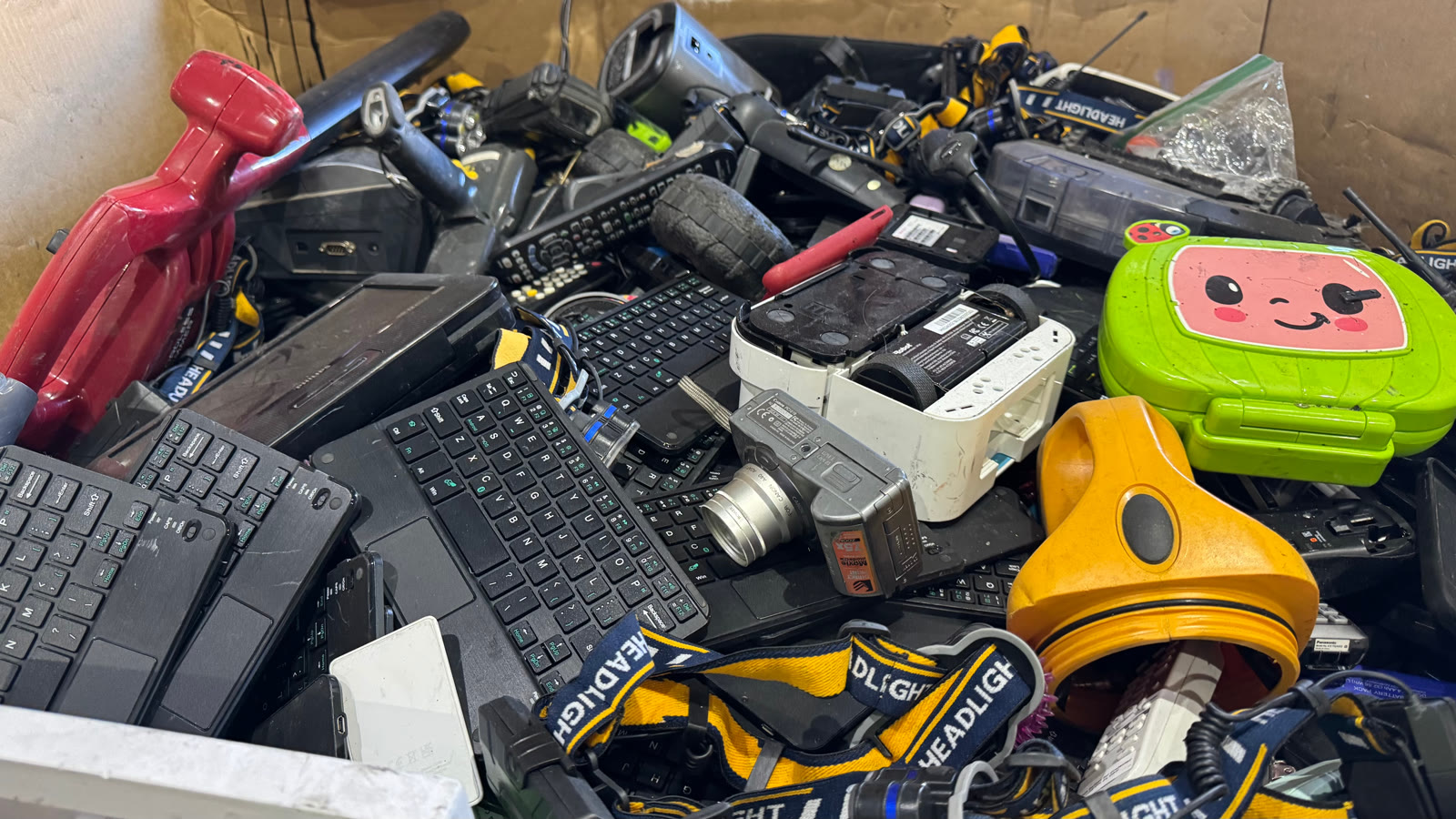 A box of small electronics for recycling: headlamps, Bluetooth keyboards, TV remotes, a camera and a toy laptop